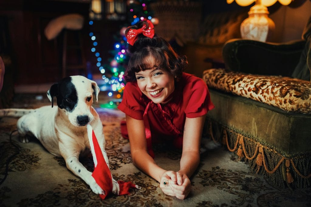 Smiling woman in red with her dog celebrating Christmas indoors with festive lights.